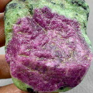 May include: A natural gemstone with a striking magenta core encased in a speckled green and black outer layer. The stone has a textured, crystalline surface. This mineral specimen is suitable for collectors and enthusiasts.