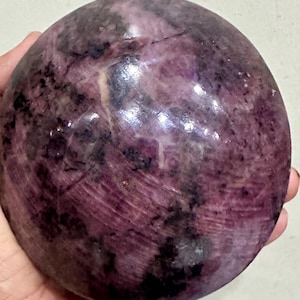Puede incluir: Una piedra esférica pulida que presenta una paleta de colores púrpura y magenta intensos con inclusiones oscuras y moteadas, además de vetas naturales visibles en toda la superficie. El objeto tiene un acabado liso, brillante y circular.