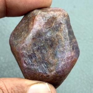 Puede incluir: Una gema de rubí en bruto y sin cortar, con una forma natural e irregular. La piedra muestra una mezcla de tonos rojo oscuro, marrón y púrpura, con estructuras cristalinas visibles. La superficie tiene un aspecto ligeramente pulido.