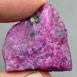 May include: A rough, uncut ruby gemstone. The ruby is a vibrant pinkish-red color with a textured surface. The gemstone has a natural, irregular shape, with visible mineral inclusions and variations in color.