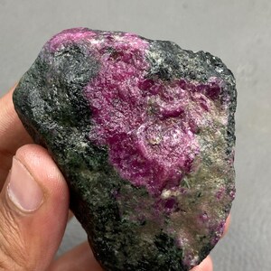 Könnte beinhalten: Ein roher, ungeschliffener Rubin-Edelstein mit einem lebendigen, rötlich-rosa Farbton. Der Stein ist teilweise in einer dunklen, gesprenkelten Matrix eingeschlossen, was einen auffälligen Kontrast erzeugt. Die rohe, natürliche Form des Rubins ist sichtbar und zeigt seine einzigartige Textur und Farbvariationen.