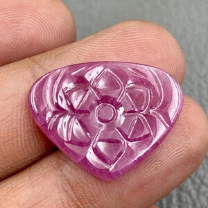 May include: A heart-shaped, pink gemstone with a carved floral design. The gemstone has a central circle with surrounding petals and geometric patterns. The polished stone is held in a hand, highlighting its intricate details. The gemstone is in a vibrant pink colour.