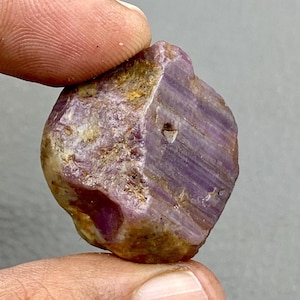 May include: A natural ruby gemstone, exhibiting a raw, unpolished appearance. The stone's surface shows a blend of purple and brown colours, with visible crystalline patterns. The ruby is held between fingers, highlighting its irregular shape and texture.