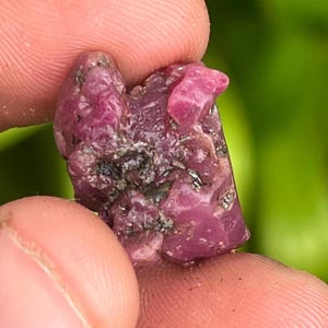 May include: A close-up of a pink ruby crystal with a rough, natural texture. The crystal has a deep pink colour and is surrounded by a dark grey matrix.