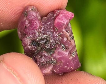 May include: A close-up of a pink ruby crystal with a rough, natural texture. The crystal has a deep pink color and is surrounded by a dark gray matrix.