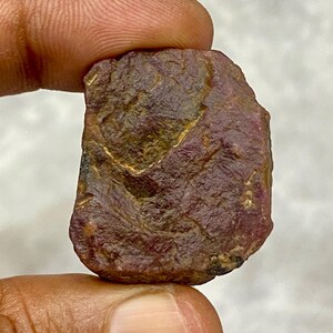 May include: A raw, uncut ruby gemstone held between fingers. The ruby is a deep red colour with brown and gold areas, and a textured surface. The gemstone is irregularly shaped and appears natural.