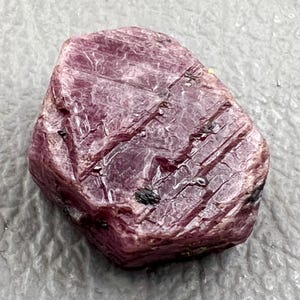 May include: A rough-cut, natural ruby gemstone. The ruby is a deep, rich pinkish-red color with visible striations and a slightly uneven surface. Small dark inclusions are present on the surface.