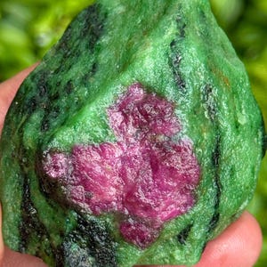 Op de afbeelding: Een ruw geslepen edelsteen met een levendige kleurencombinatie. De steen heeft een diepgroene basis met opvallende zwarte aderen. Een prominente, onregelmatige vlek van levendig magenta zorgt voor een gedurfd contrast, waardoor een visueel boeiend mineraal specimen ontstaat.