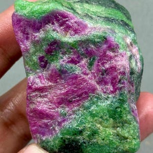 Peut inclure: Une pierre précieuse brute de rubis zoïsite. Cette gemme présente un mélange de couleurs vives, notamment du magenta profond, du vert vif et du noir. La texture rugueuse et la forme naturelle de la pierre sont visibles, mettant en valeur sa beauté.