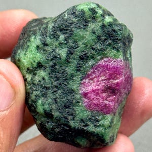 May include: A rough ruby in matrix, held in a hand. The stone has a vibrant magenta centre surrounded by green and black mottled areas. The natural gemstone has a textured surface.
