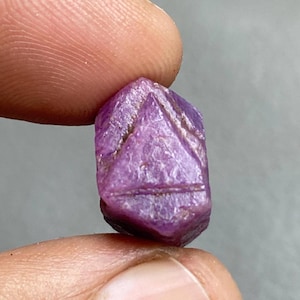 May include: A small, faceted, purplish-pink gemstone held between fingers. The gem has a geometric design carved into its surface. The stone's color is a vibrant, saturated pink, with a slightly rough texture. The background is a neutral gray.