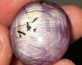 Pode incluir: Uma gema redonda de cor púrpura com uma superfície lisa e polida. A pedra tem um brilho iridescente leve e é coberta por linhas finas e pretas.