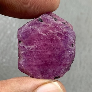 May include: A rough, uncut ruby gemstone, held between fingers. The ruby is a deep pinkish-red colour with a textured surface and small black inclusions. The gemstone has an irregular, faceted shape, showcasing its natural form.