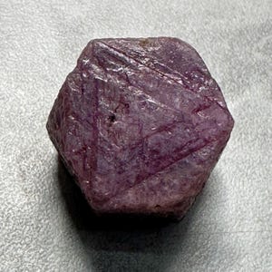 May include: A rough, faceted ruby gemstone with a deep reddish-purple hue. The natural stone has a hexagonal shape with visible surface striations and a slightly uneven texture. The gem is displayed against a neutral background.