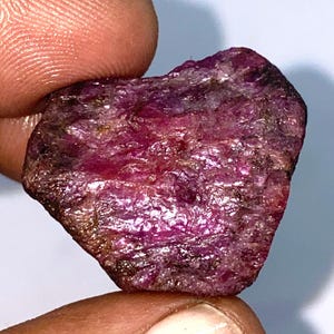 May include: A rough, heart-shaped ruby gemstone with a deep pink hue. The gemstone has a natural, unpolished finish and shows intricate striations.