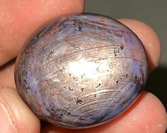 Pode incluir: Uma gema lisa, ovalada, com um brilho iridescente roxo e prateado. A pedra tem um padrão estriado e inclusões pretas.