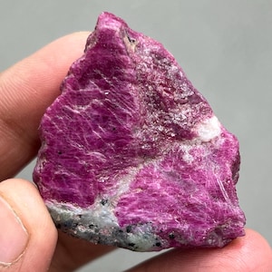 Pode incluir: Um cristal de rubi rosa bruto com uma matriz cinza escura e branca. O cristal tem uma cor rosa profunda e rica e é translúcido.