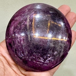 Puede incluir: Una piedra pulida de forma circular con una paleta de colores magenta intenso y morado oscuro. La superficie presenta una textura lisa y reflectante con inclusiones naturales visibles y patrones cristalinos internos en toda la gema.