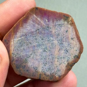 May include: A natural gemstone with a mix of colours, including deep reds, purples, and browns. The stone has a natural, unpolished appearance, with visible lines and textures across its surface. The overall shape is irregular, with a slightly hexagonal form.