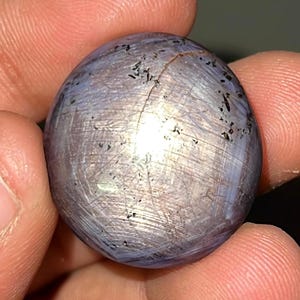 May include: A round, polished, gray gemstone with a star-like pattern and black inclusions.