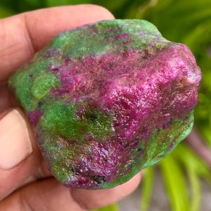 Può includere: Una gemma di rubino grezzo in zoisite. La pietra presenta una vivace miscela di colori, con una profonda tonalità magenta che domina un lato e un verde intenso sull'altro. La superficie ha una texture naturale e non lucidata.