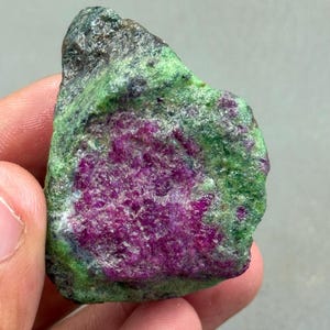 May include: A raw gemstone with a striking colour palette. The stone showcases a rich magenta core, enveloped by a speckled green and dark grey outer layer. The natural textures and colour variations give it a unique appearance.