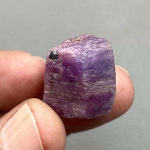 May include: A raw, uncut ruby gemstone, held between fingers. The ruby is a deep purple colour with a rough, textured surface and a small black inclusion. The gemstone is a natural mineral specimen.