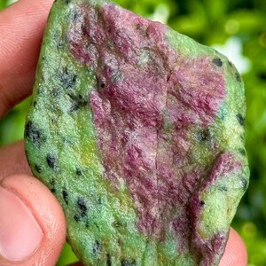 Peut inclure: Une pierre précieuse brute avec une palette de couleurs vibrantes. La pierre présente un mélange de vert, de rouge profond et de noir. La surface a un aspect texturé, avec les couleurs qui se fondent. La pierre est tenue dans une main, avec un arrière-plan vert flou.