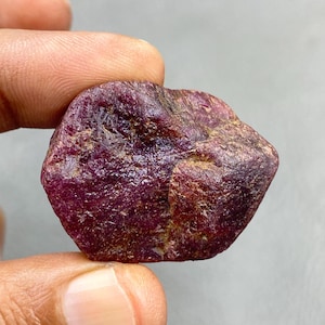 Può includere: Una gemma di rubino grezza, non tagliata, di colore rosso intenso. La superficie della pietra ha un aspetto strutturato con aree di tonalità rosse e marroni più chiare e scure. Il rubino è tenuto tra due dita su uno sfondo neutro.