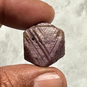May include: A raw ruby gemstone, held between fingers. The ruby is a deep reddish-purple colour with etched geometric patterns on its surface. The gemstone is held against a neutral background.