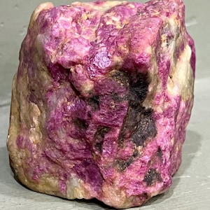Könnte beinhalten: Ein rohes, unpoliertes Mineralexemplar mit einer leuchtend magentafarbenen und tiefrosa kristallinen Struktur. Der Stein weist eine raue, texturierte Oberfläche mit eingestreuten Flecken aus undurchsichtigen weißen und dunkelgrauen Einschlüssen auf.