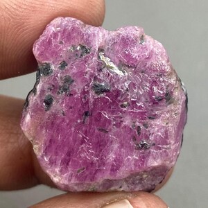 Op de afbeelding: Een ruwe, ongeslepen robijn edelsteen. De robijn heeft een diep roze-rode kleur met donkergrijze en zwarte insluitsels. Het oppervlak van de steen is ongelijkmatig en getextureerd, wat de natuurlijke, ruwe vorm benadrukt. Een mooi voorbeeld van natuurlijke minerale formaties.