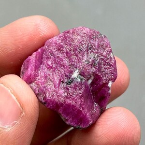 May include: A rough, pink ruby crystal with a dark grey inclusion. The crystal is approximately 5 cm long and 4 cm wide.