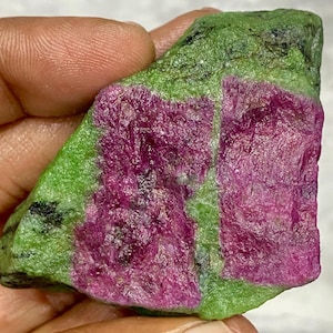 Può includere: Una gemma grezza con una vibrante combinazione di colori. La pietra mostra un contrasto sorprendente tra il magenta intenso delle inclusioni di rubino e la zoisite verde smeraldo circostante. La texture naturale e le variazioni di colore creano un aspetto unico.