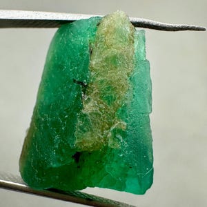 May include: A raw emerald gemstone, held by tweezers. The emerald is a vibrant green color with a rough, textured surface. A section of the stone has a lighter, yellowish-green hue. The gemstone is irregularly shaped and appears to be uncut.