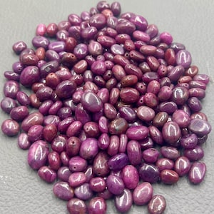 Puede incluir: Un montón grande de pequeñas cuentas de piedras preciosas ovaladas de color púrpura. Las cuentas son lisas y brillantes, y tienen una apariencia ligeramente translúcida.
