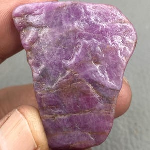 Op de afbeelding: Een ruw geslepen, natuurlijke robijn edelsteen met een diepe roze-paarse tint. De steen heeft een getextureerd oppervlak met zichtbare minerale patronen en kleurvariaties, variërend van lichte tot donkere roze tinten.