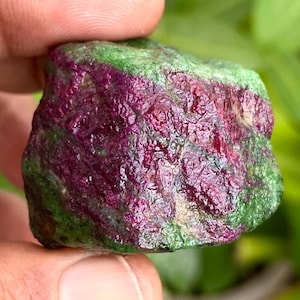 Peut inclure: Une pierre précieuse brute de rubis-en-zoïsite. La pierre présente un aspect moucheté avec un mélange de couleurs magenta foncé et vert forêt. La surface a une texture légèrement rugueuse, avec des structures cristallines visibles.
