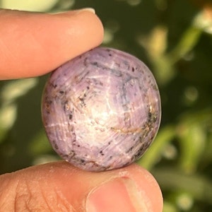 May include: A round, polished gemstone with a light purple hue, held between fingers. The stone has a star-like pattern and dark inclusions. The background is a blurred green.