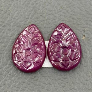 Puede incluir: Dos gemas de rubí talladas, cada una en forma de lágrima. Las gemas tienen un diseño floral tallado en su superficie. El color es un rojo intenso y rico. Las gemas se muestran sobre una superficie gris.