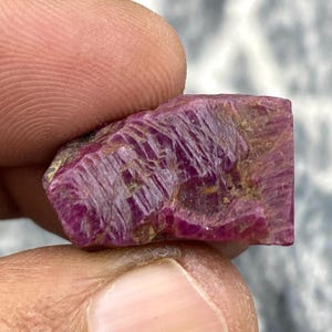 May include: A raw ruby gemstone, held between fingers, presents a deep pinkish-red colour with visible crystalline structures. The unpolished mineral has a textured surface, showcasing its natural beauty. The ruby is a rough-cut specimen.