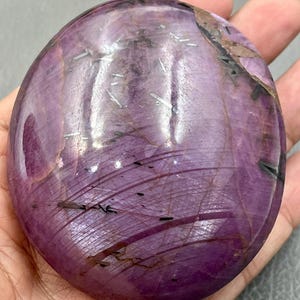 May include: A polished, oval-shaped purple gemstone featuring a smooth, glossy surface with visible internal inclusions, dark linear streaks, and subtle white crystalline patterns throughout the stone's translucent and opaque violet body.