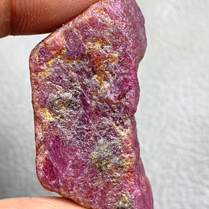 May include: A rough, uncut ruby gemstone with a deep red hue. The surface has a textured appearance with areas of gold and black coloration. The ruby is held between fingers, showcasing its natural, raw form. This gemstone is ideal for jewelry making or collecting.