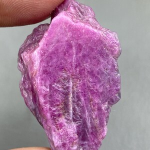 Könnte beinhalten: Ein rauer, ungeschliffener Rubin-Edelstein mit einem tiefen, rötlich-rosa Farbton. Der Naturstein hat eine strukturierte Oberfläche mit sichtbaren kristallinen Strukturen und Farbintensitätsvariationen. Der Edelstein wird zwischen zwei Fingern gehalten.