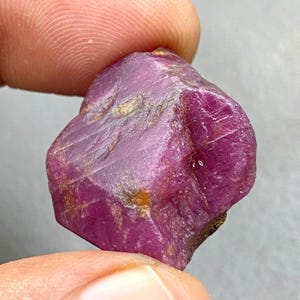 May include: A rough, uncut ruby gemstone with a deep pinkish-red hue. The natural stone has an irregular shape with visible surface texture and small areas of lighter color. The ruby is held between two fingers.