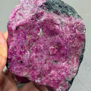 Puede incluir: Una gema de rubí en bruto, con un vibrante tono rosa-rojizo. La piedra tiene una superficie texturizada con áreas negras y grises, mostrando su estado bruto y sin pulir. Un espécimen mineral único para coleccionistas.