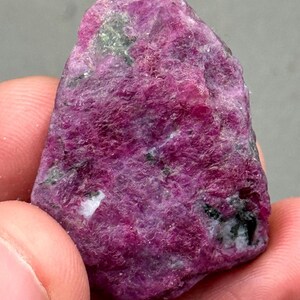 Puede incluir: Una gema de rubí en bruto sin cortar. El rubí es de un rojo intenso y vibrante con áreas de rosa más claro e inclusiones de color verde oscuro. La superficie tiene un aspecto texturizado e irregular, mostrando su forma natural y en bruto. La gema se sostiene en una mano.