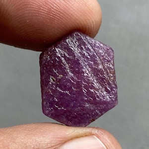 May include: A rough-cut, hexagonal ruby gemstone. The ruby is a deep, saturated purple color with a textured surface. The gemstone is held between two fingers against a neutral background.
