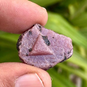 Può includere: Un cristallo di rubino rosa con forma triangolare e inclusioni nere. Il cristallo è grezzo e ha un aspetto naturale non lucidato.