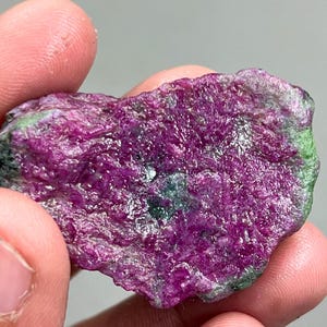 Puede incluir: Una gema áspera de color rosa y verde con una forma natural irregular. La gema tiene una superficie brillante y pulida.